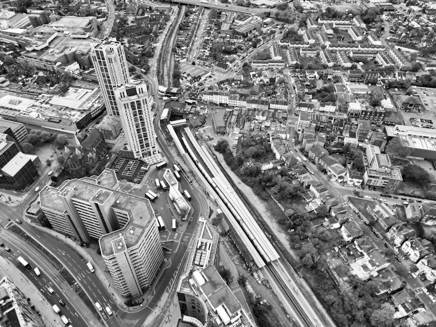An aerial black-and-white photograph of a dense urban area showing high-rise office buildings, residential structures, and tightly packed houses with pitched roofs, roads, and trees. A railway line with multiple train carriages runs through the middle of the scene, parallel to a street where several buses and cars are visible. The surrounding streets are busy with vehicular traffic, and the area includes a mix of commercial and residential zones. The overall environment is typical of a central business and residential district, with complex street layouts and varying building heights. This type of urban landscape reflects the city infrastructure that a professional removal service like Man and Van Colliers Wood may operate within when providing house removals and furniture transport services near Colliers Wood Station.