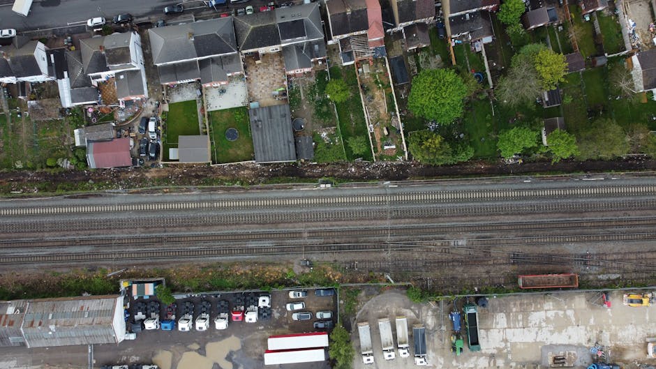 An aerial black-and-white photograph of a dense urban area showing high-rise office buildings, residential structures, and tightly packed houses with pitched roofs, roads, and trees. A railway line with multiple train carriages runs through the middle of the scene, parallel to a street where several buses and cars are visible. The surrounding streets are busy with vehicular traffic, and the area includes a mix of commercial and residential zones. The overall environment is typical of a central business and residential district, with complex street layouts and varying building heights. This type of urban landscape reflects the city infrastructure that a professional removal service like Man and Van Colliers Wood may operate within when providing house removals and furniture transport services near Colliers Wood Station.
