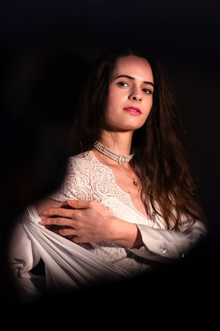 A woman with long, dark hair and fair skin is seen indoors against a dark background. She is wearing a white, lace-textured top or dress with a high neckline, and accessorized with a multi-strand pear