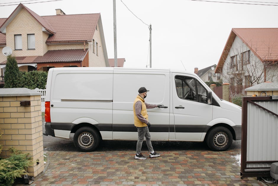 A man wearing a face mask, glasses, a beige and brown jacket, and dark grey trousers is seen standing on a brick driveway, opening the sliding door of a white cargo van for a home relocation. The van is parked on the driveway of a residential property situated in a suburban area with semi-detached houses featuring pitched roofs and brick walls. The van is a medium-sized commercial vehicle with black wheel trims and a grey lower trim, positioned parallel to a low brick wall and a grey metal gate on either side. The driveway surface consists of multicoloured paving bricks, and the background includes houses with red-tiled roofs, windows, and a utility pole with overhead electrical wires. The scene suggests a professional removals or furniture transport process, likely carried out by [COMPANY_NAME], as part of packing and moving services for High Street flats in Colliers Wood, SW19.