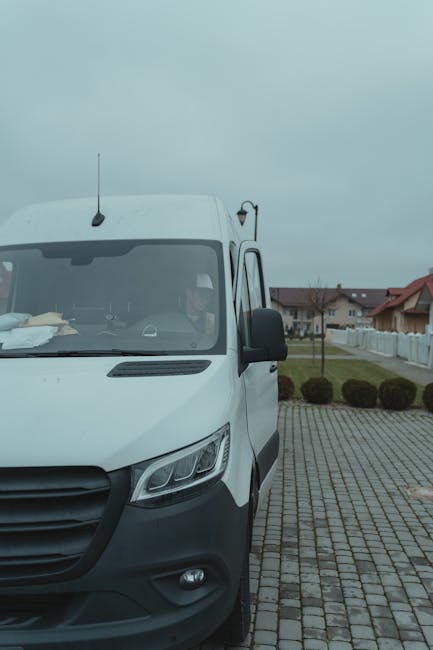A white van parked on a cobblestone driveway outside a residential building under an overcast sky. The front of the van is visible, with the driver seated inside, partially obscured by the windshield. The van features black side mirrors, an antenna on the roof, and sleek headlights. Inside the van, packing materials such as cardboard boxes and plastic wrap are visible on the dashboard, indicating a home relocation process coordinated by Man and Van Colliers Wood. The driveway area is bordered by a low white fence, with neatly trimmed bushes and neighboring houses in the background. The scene captures the preparation stage of furniture transport and packing for a move within the High Street flats area in Colliers Wood, illustrating the logistics involved in professional removals.