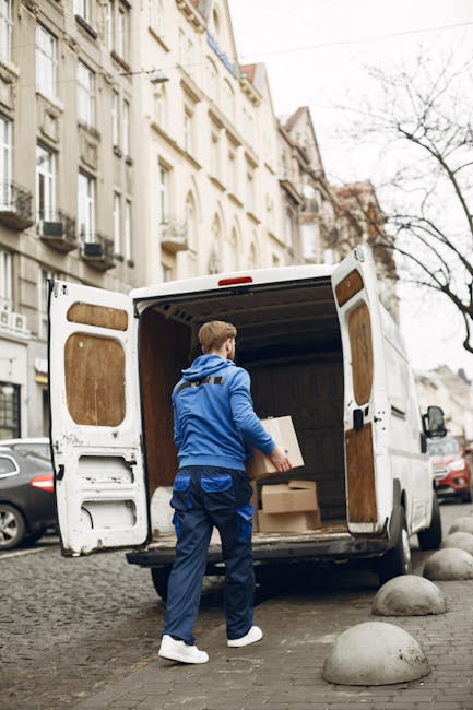 A man wearing a face mask, glasses, a beige and brown jacket, and dark grey trousers is seen standing on a brick driveway, opening the sliding door of a white cargo van for a home relocation. The van is parked on the driveway of a residential property situated in a suburban area with semi-detached houses featuring pitched roofs and brick walls. The van is a medium-sized commercial vehicle with black wheel trims and a grey lower trim, positioned parallel to a low brick wall and a grey metal gate on either side. The driveway surface consists of multicoloured paving bricks, and the background includes houses with red-tiled roofs, windows, and a utility pole with overhead electrical wires. The scene suggests a professional removals or furniture transport process, likely carried out by [COMPANY_NAME], as part of packing and moving services for High Street flats in Colliers Wood, SW19.